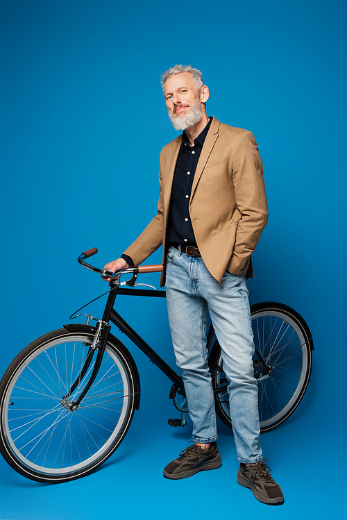 full length of pleased middle aged man standing with hand in pocket near bicycle on blue