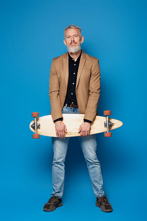 full length of bearded middle aged man with grey hair standing with longboard on blue