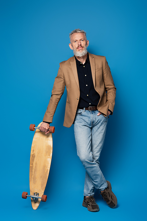 full length of bearded middle aged man standing with hand in pocket near longboard on blue
