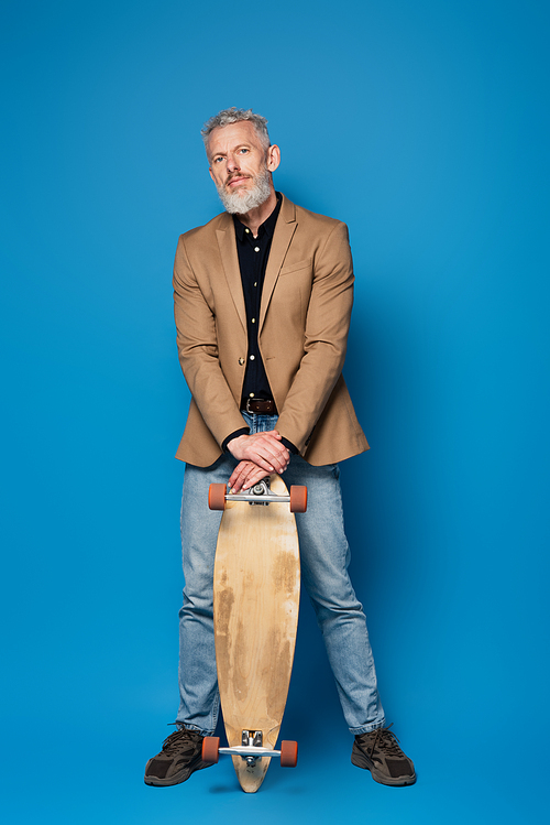 full length of bearded middle aged man standing with longboard on blue