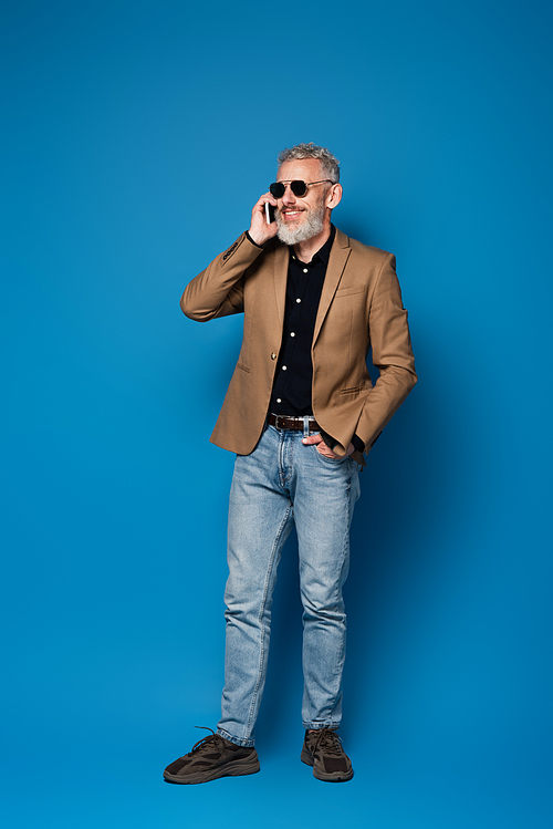 full length of happy middle aged man in sunglasses talking on cellphone on blue