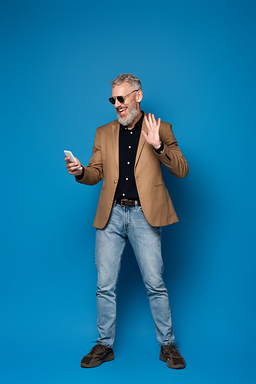 full length of happy middle aged man in sunglasses having video call on cellphone and waving hand on blue