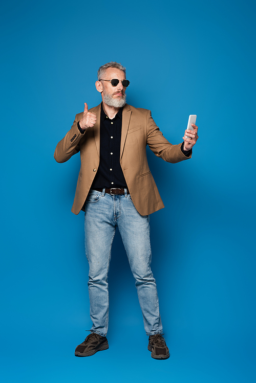 full length of middle aged man in sunglasses showing thumb up while taking selfie on blue