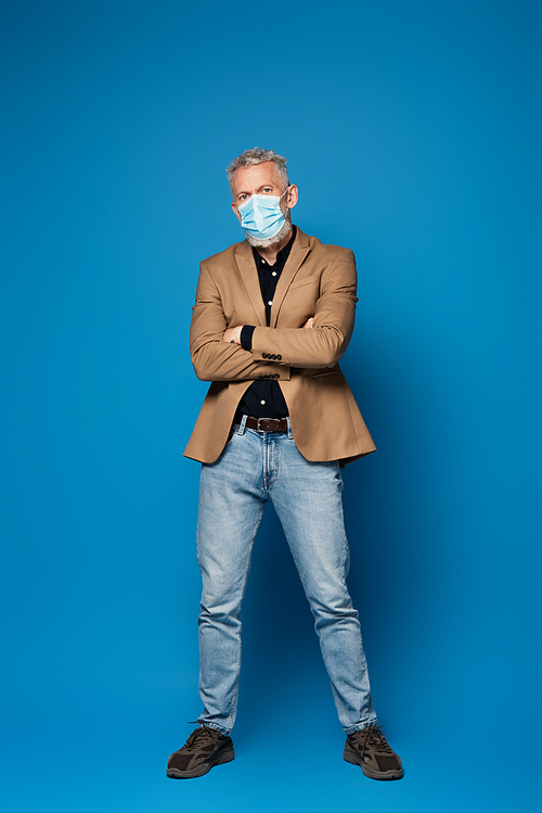 full length of middle aged man in protective medical mask standing with crossed arms on blue