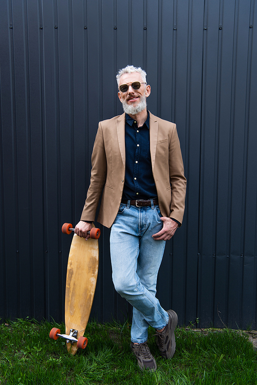 full length of cheerful middle aged man in sunglasses holding longboard outside