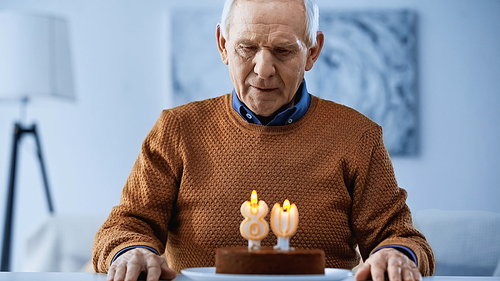 lonely elderly man looking at burning candles on birthday cake in living room