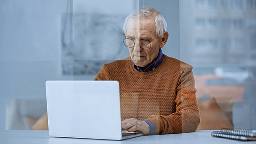 serious elderly man sitting at table near window and working on laptop at home