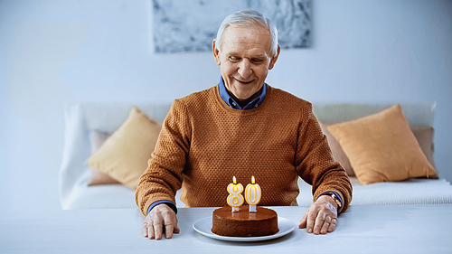 happy elderly man celebrating birthday in front of cake with burning candles in living room