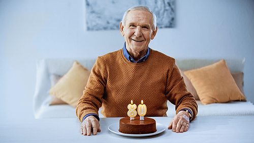 cheerful elderly man sitting in front of birthday cake with burning candles in modern living room