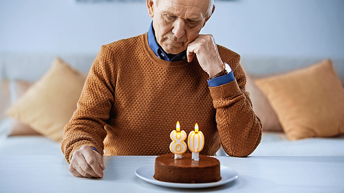 sad elderly man sitting alone in front of birthday cake with candles in living room