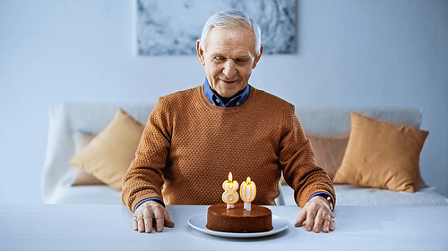 happy elderly man sitting in front of birthday cake with candles in living room