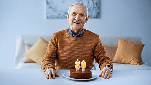 happy elderly man sitting alone in front of birthday cake with candles in living room