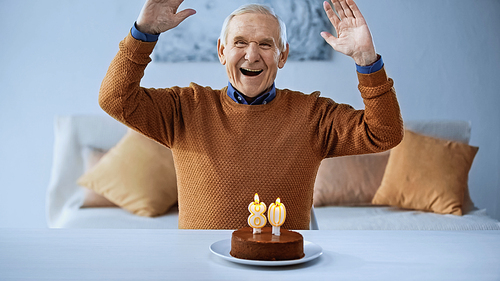 excited elderly man celebrating birthday in front of cake with burning candles in living room