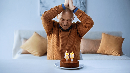 cheerful elderly man sitting in front of birthday cake with burning candles in living room