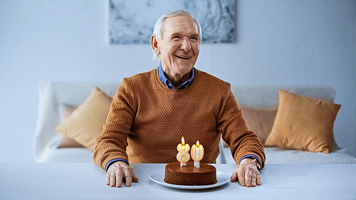 happy elderly man celebrating birthday in front of cake with burning candles in living room in living room