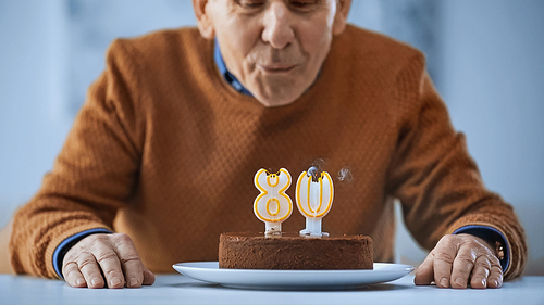 cheerful elderly man blowing out candles on birthday cake on grey background
