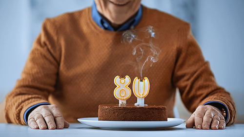 cropped view of elderly man celebrating birthday in front of cake with blown out candles on grey background