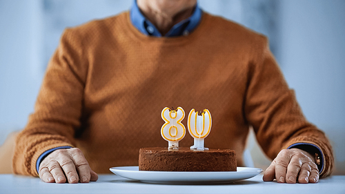 cropped view of elderly man sitting in front of birthday cake with blown out candles on grey background