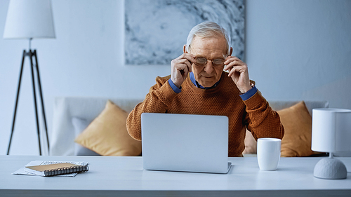 elderly man adjusting glasses while working near laptop in living room