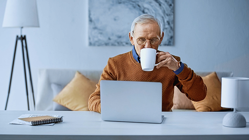 elderly man drinking coffee and working from home with laptop in living room