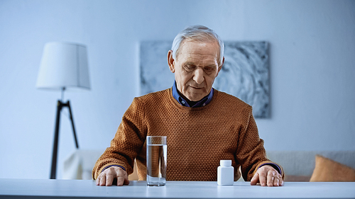 elderly man sitting near table with medicine jar and glass of water in living room