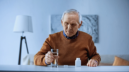elderly man sitting with open medicine jar on table and holding glass of water in living room