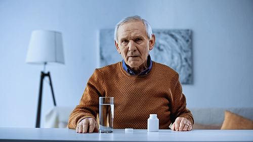 elderly man sitting with medicine jar and glass of water in living room