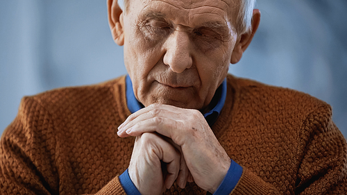 portrait of elderly man with closed eyes and clenched hands near face on grey background