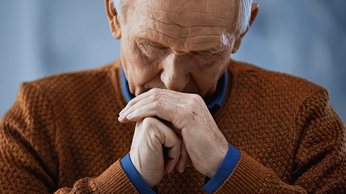 portrait of elderly man with clenched hands near face and closed eyes on grey background