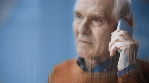 sad elderly man looking outside through rainy window and speaking on cellphone grey background behind rainy glass