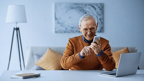smiling elderly man sitting near laptop and messaging on smartphone at home