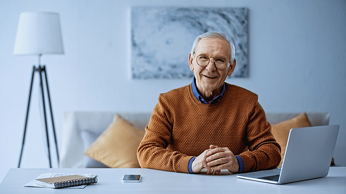 happy elderly man sitting with clenched hands at table with laptop and cellphone in living room