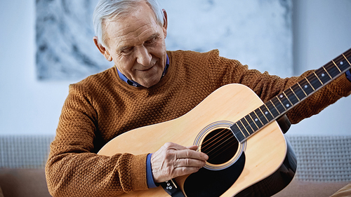 smiling elderly man sitting on sofa and playing guitar in living room