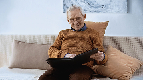 smiling elderly man sitting on sofa with family album at home