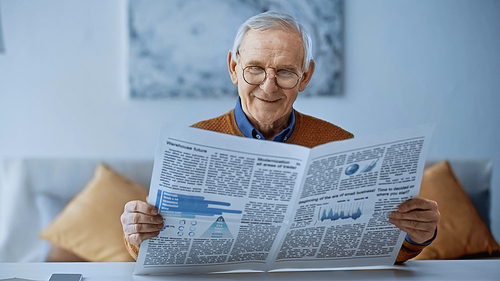 smiling elderly man in glasses reading newspaper at home