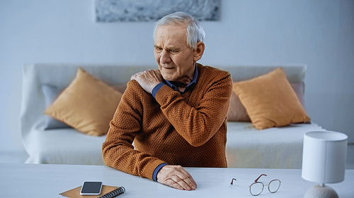 suffering elderly man sitting at table and touching his shoulder at home