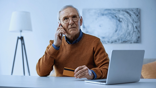 serious elderly man sitting with credit card near laptop and talking on cellphone at home