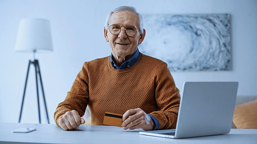 happy elderly man in glasses sitting near laptop and cellphone and holding credit card in living room