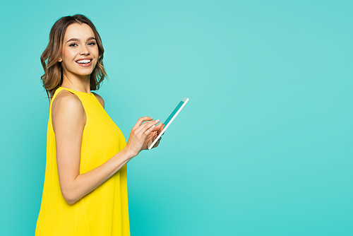 Woman in dress using digital tablet and smiling at camera isolated on blue