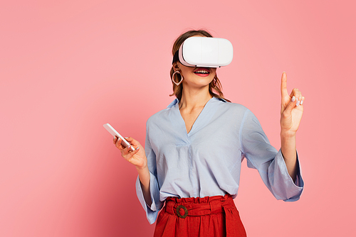 Smiling woman in vr headset pointing with finger on pink background