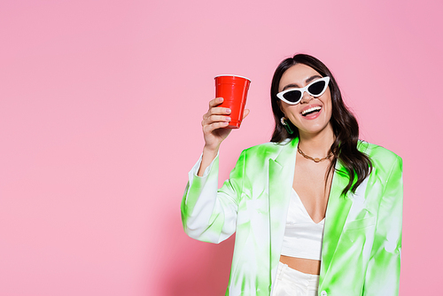 Cheerful woman in jacket and sunglasses holding plastic cup on pink background