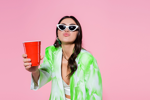 Stylish woman in sunglasses holding plastic cup and pouting lips isolated on pink