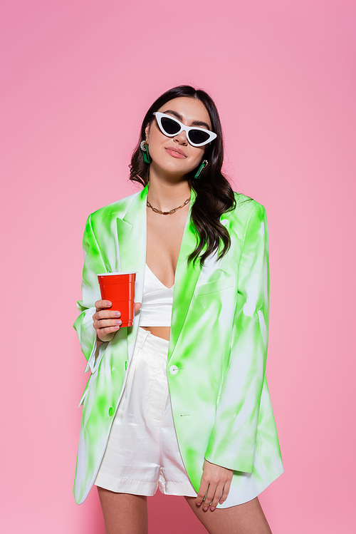 Young woman in blazer and sunglasses holding plastic cup on pink background