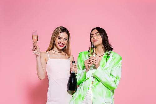 Cheerful woman holding glass and bottle with champagne near friend on pink background