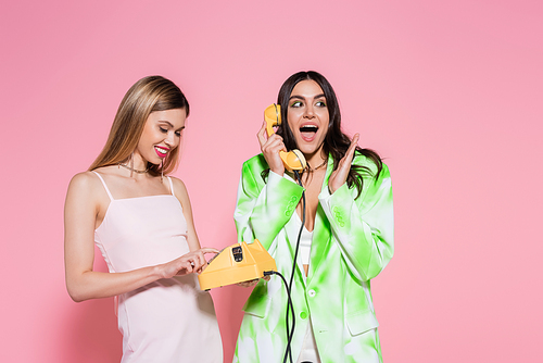 Smiling woman holding telephone near excited friend talking on pink background