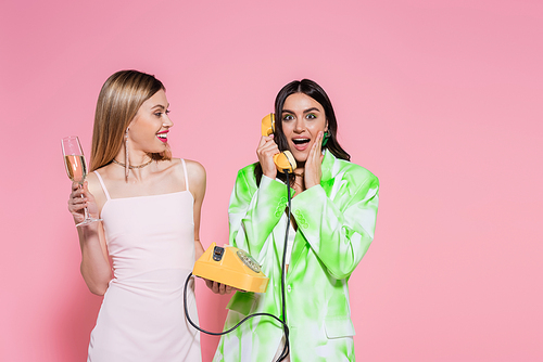 Excited woman talking on telephone near friend with champagne on pink background