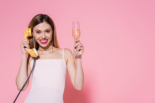 Smiling woman holding champagne and talking on telephone on pink background with copy space