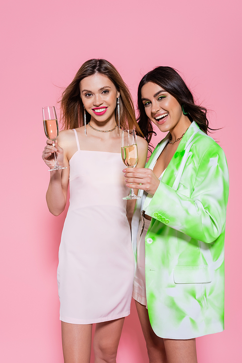 Young women in stylish clothes holding champagne on pink background