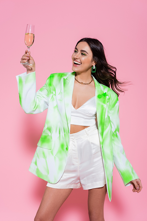 Positive woman in jacket holding glass of champagne on pink background