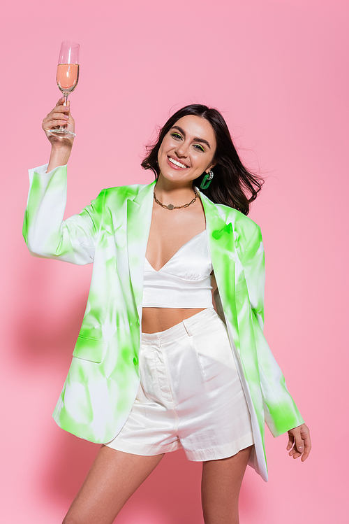 Brunette woman in stylish clothes holding champagne on pink background